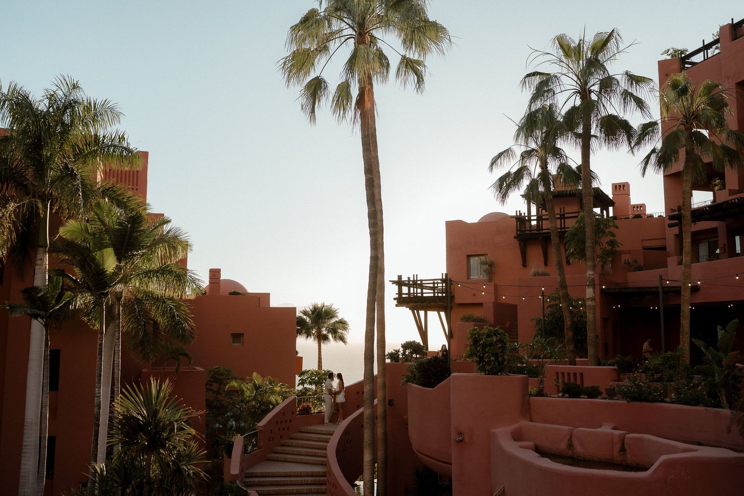 Pink terracotta resort with palm trees, curved staircases, and ocean view at golden hour in a tropical setting.