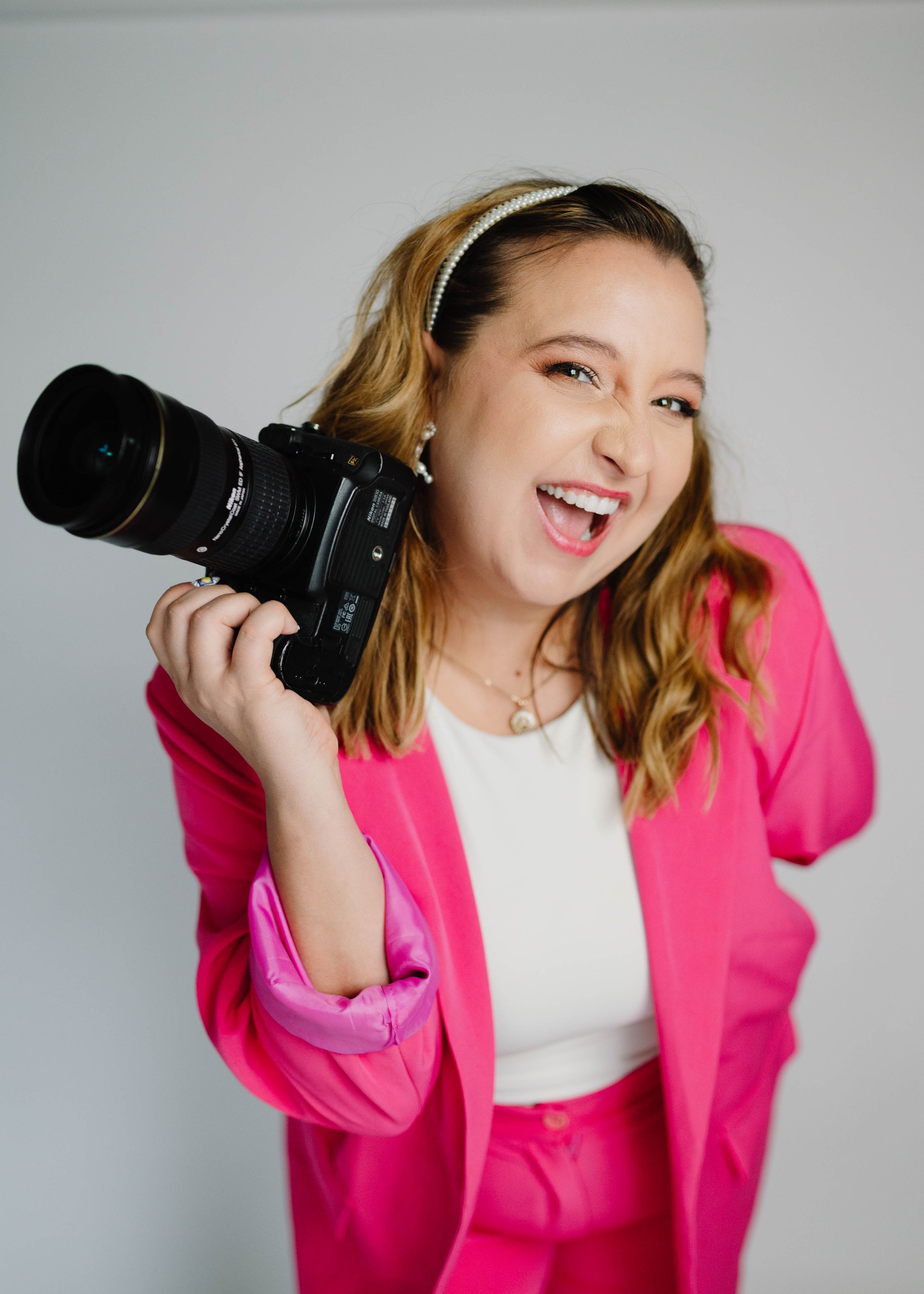 Lex in a pink power suit smiling and posing with her camera at a studio with white background. Cred: AngelaRodPhoto