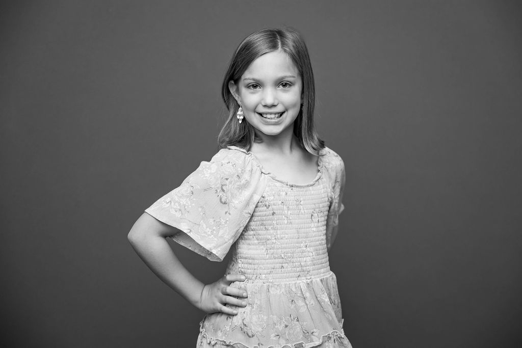 A young child in a white lace dress poses confidently against a gray studio backdrop in black and white.