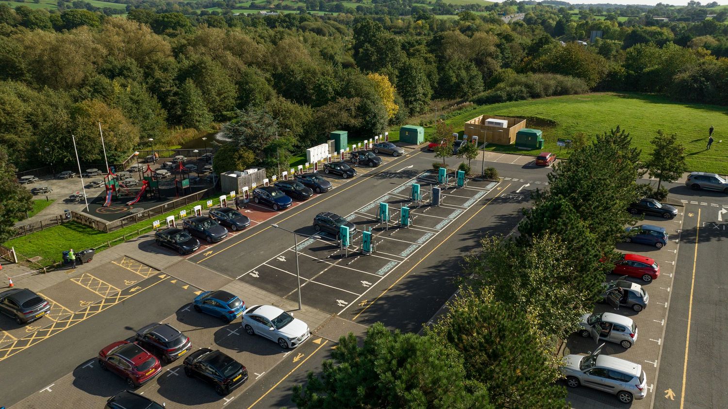 Aerial view of a parking lot with cars surrounded by green trees and countryside in bright daylight.