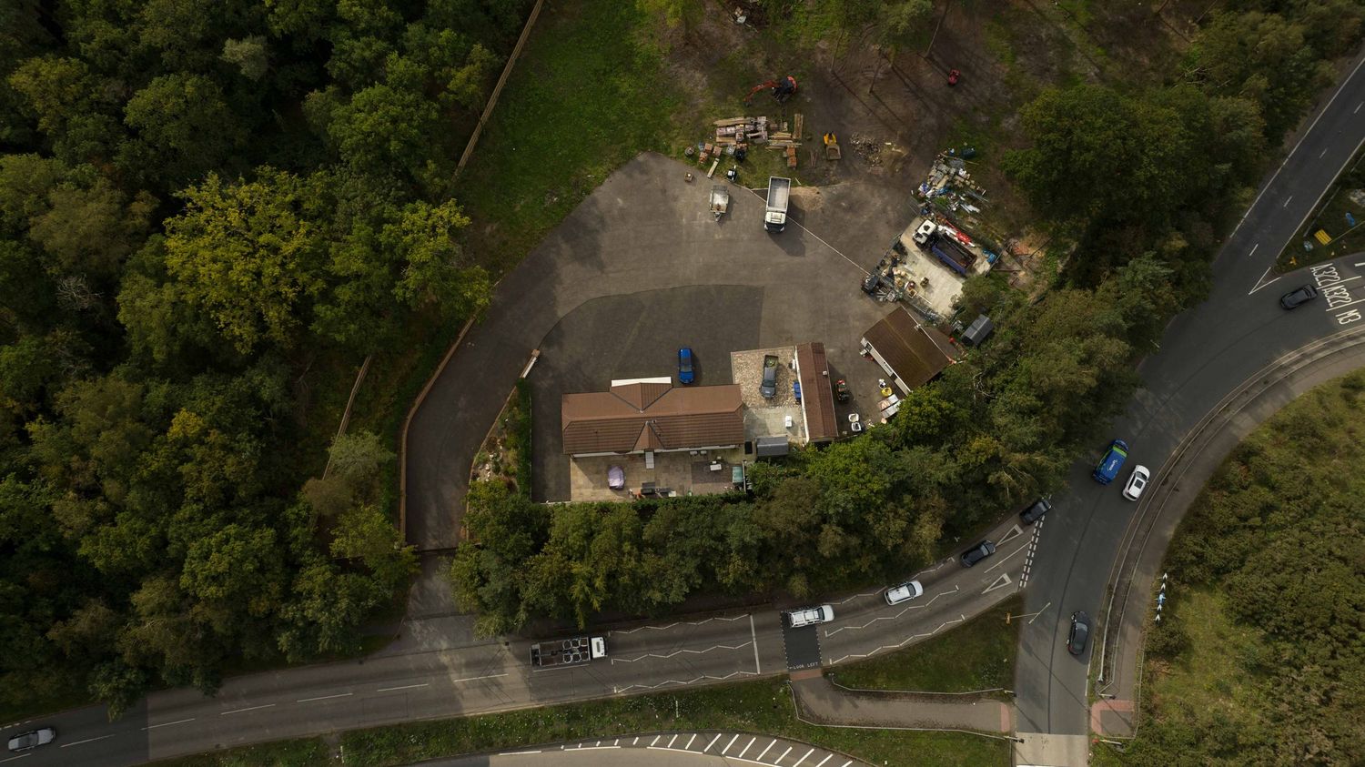 Aerial drone view of a rural house with white walls and red roof surrounded by trees and winding roads.