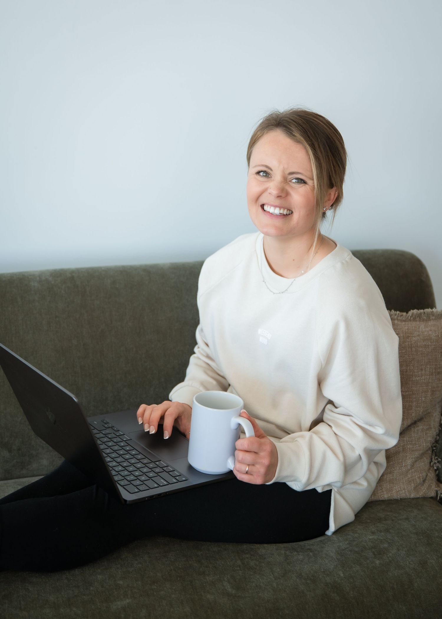 Someone works from home on a laptop while sitting on a grey couch, wearing a white sweater and holding a blue coffee mug.