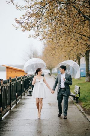 A couple walks in the rain holding clear umbrellas under golden autumn trees.