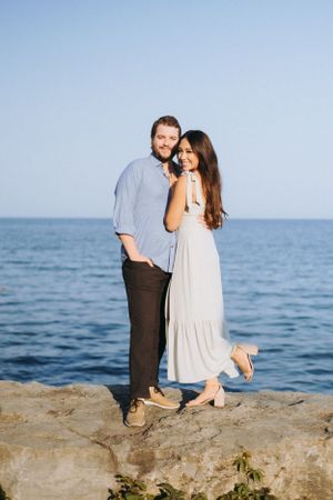 Couple poses on rocky coastline against serene blue ocean backdrop.