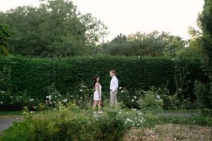 A couple stands together in a lush garden setting surrounded by hedges and flowers at dusk.