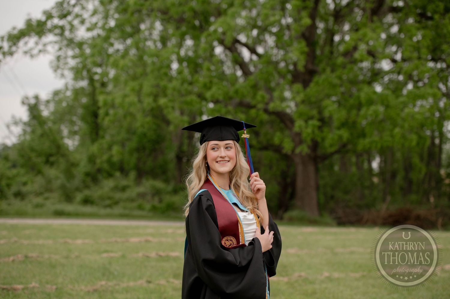 Graduate in cap and gown poses with diploma in front of green trees during outdoor commencement ceremony.