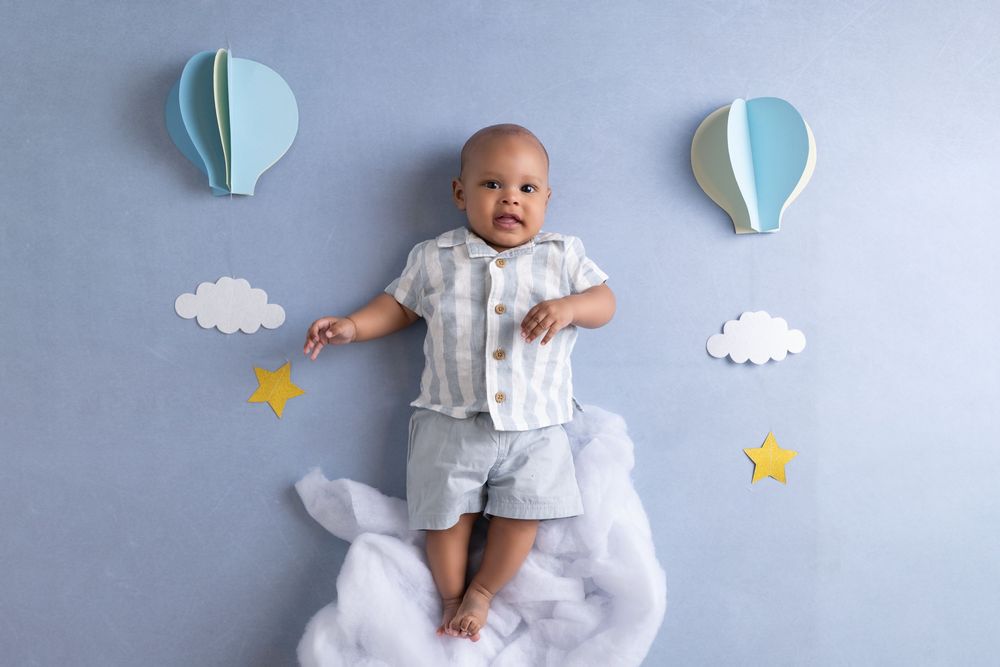 A baby in gray outfit lies on fluffy clouds with paper hot air balloons and stars on a blue background.