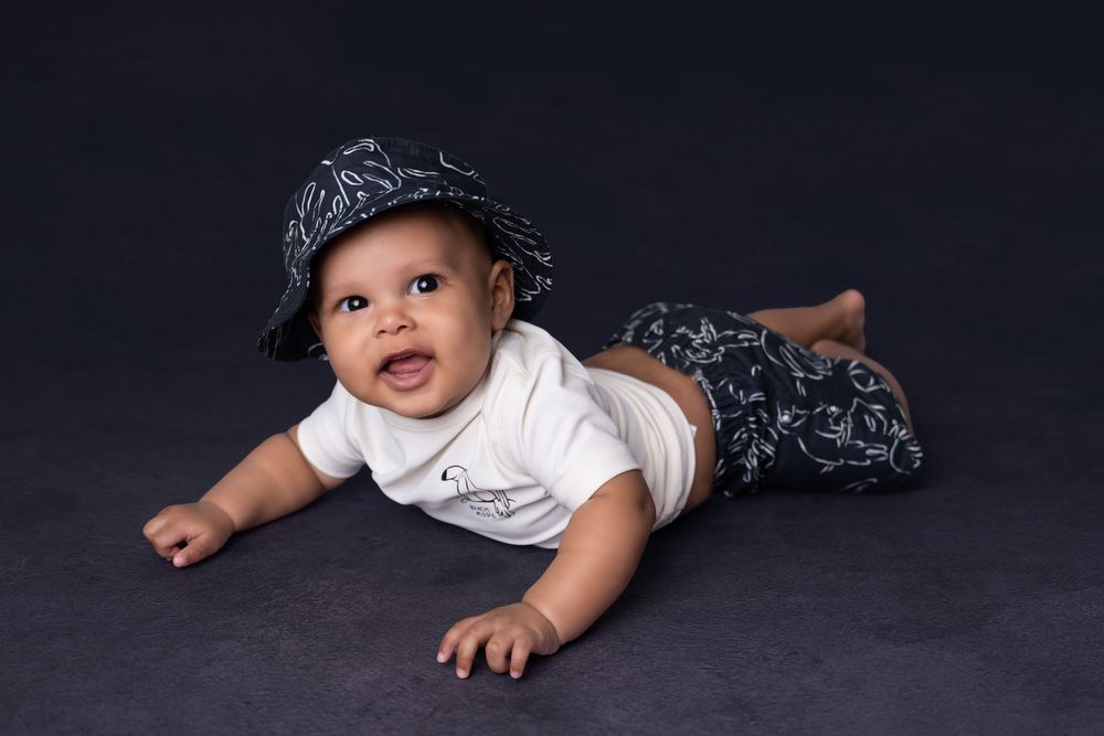 Smiling baby in a white onesie, navy bucket hat, and patterned pants lying on a dark gray studio floor.