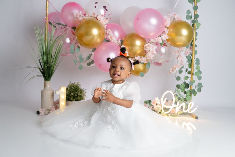 Baby girl in white dress celebrates first birthday with pink and gold balloons and "One" decorations.