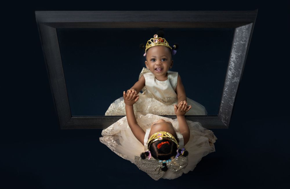 Baby girl in white dress and gold crown sits inside a wooden frame prop during a royal-themed first birthday photo session.