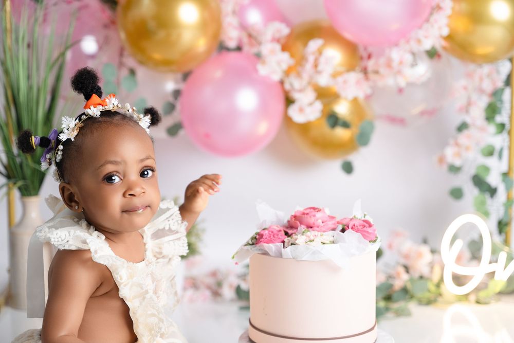 A baby girl in a white lace outfit sits beside a pink floral cake, surrounded by pink and gold balloons.