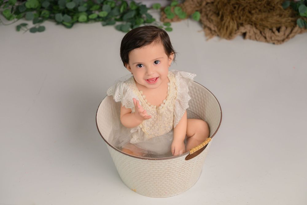 smiling baby in a cream lace romper sits inside a  white bucket during a milestone photo session milestone photographer cypress tx