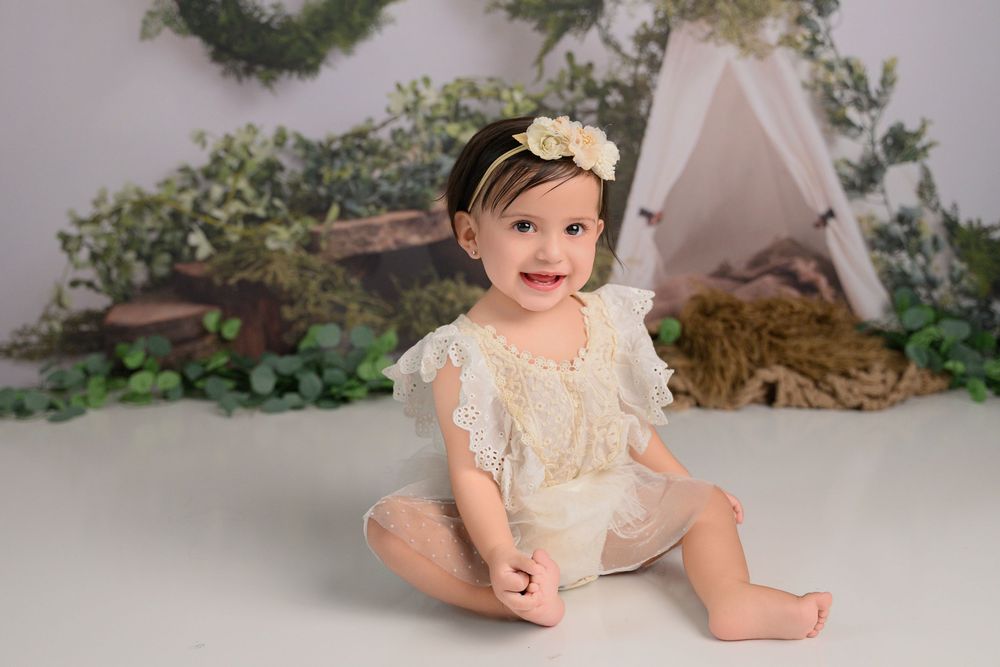 baby girl in lace romper and floral headband sits on white floor before a whimsical woodland backdrop Cypress milestone photographer