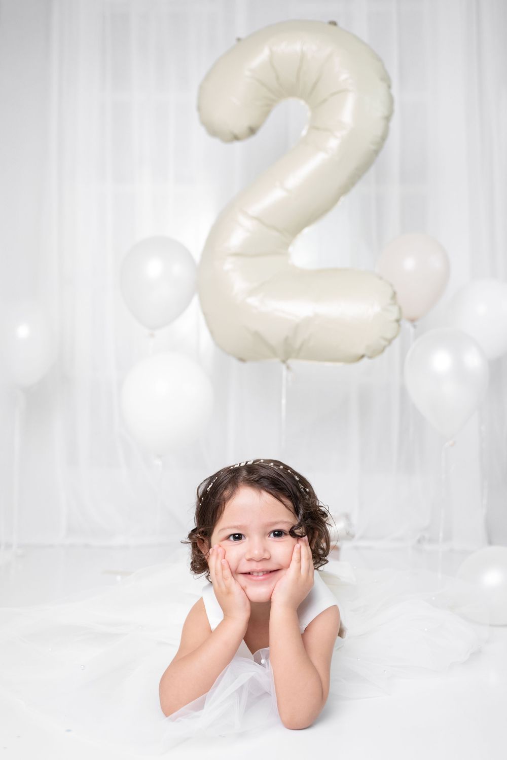 Smiling toddler girl in white dress lying down with number 2 balloon for 2nd birthday celebration. Photographer in Cypress TX