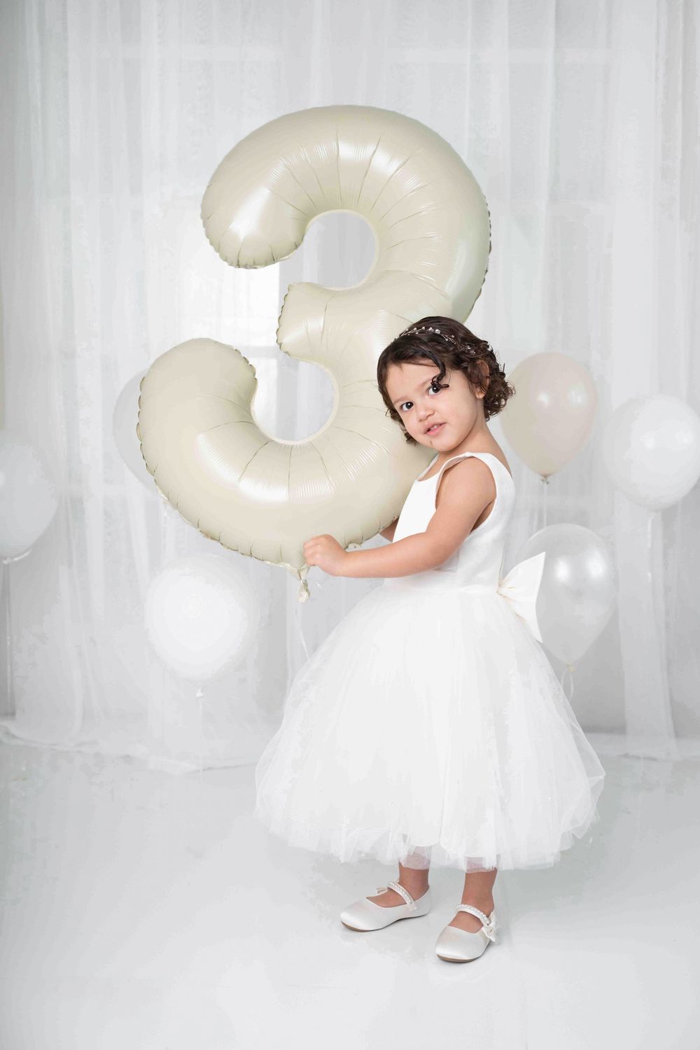 A little girl in a white tutu dress holds a large number 3 balloon at her third birthday party. Photographer in Cypress TX