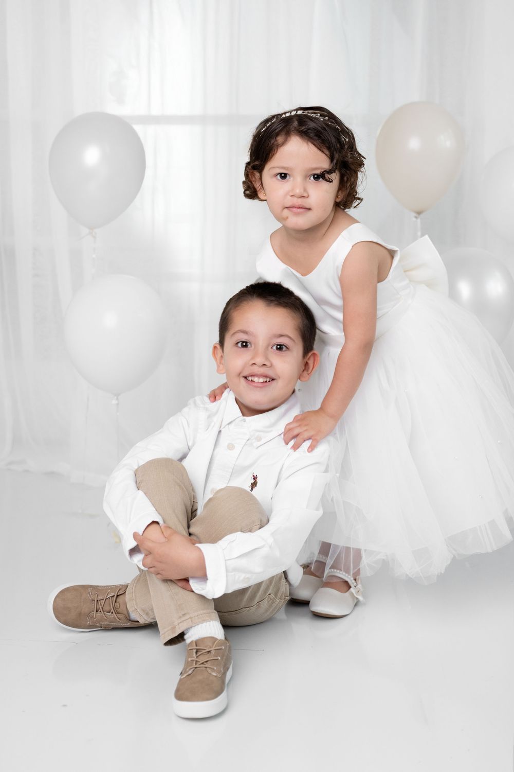 Two young children in white formal outfits pose with white balloons in a bright studio setting.