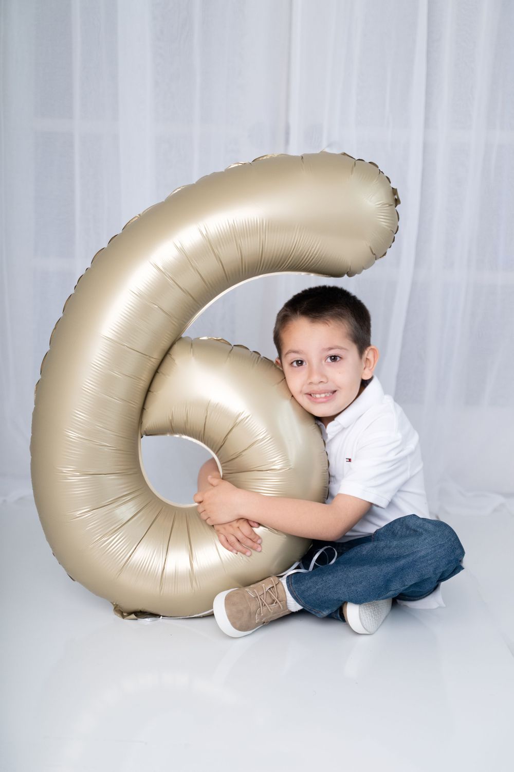 A smiling boy sits cross-legged hugging a large gold number 6 balloon against a white curtain backdrop.  Photographer in Cypress TX