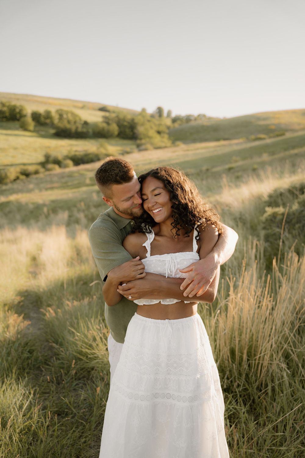 Couple shares tender embrace in tall grass during summer sunset photoshoot.