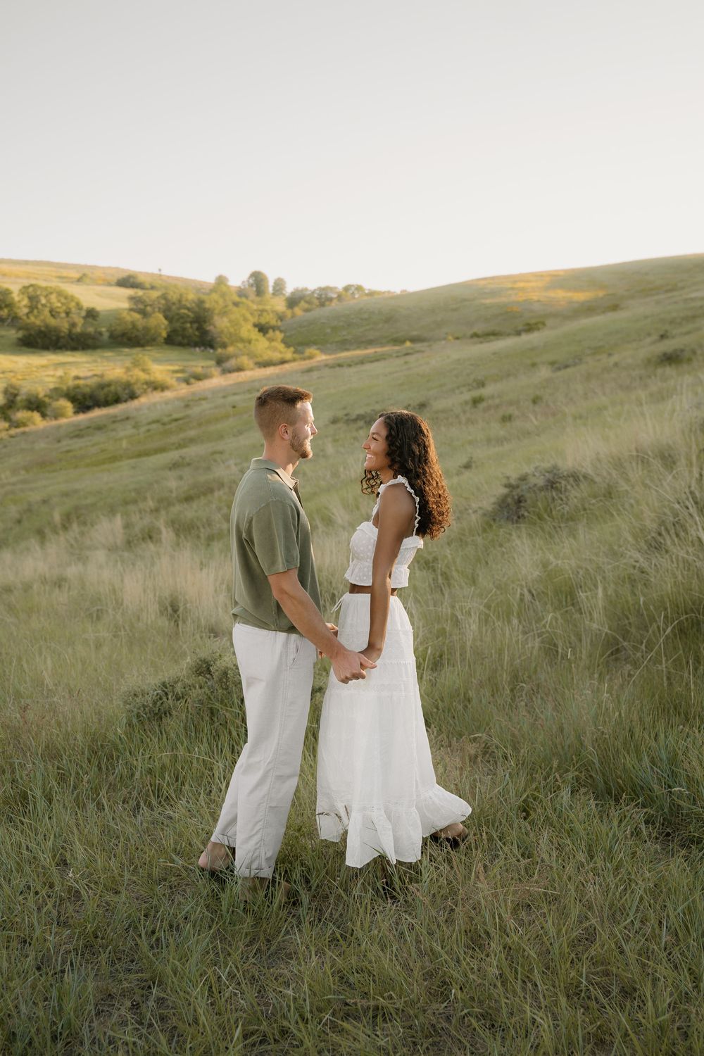 Romantic couple in coordinated white outfits stand together in rolling hillside meadow.