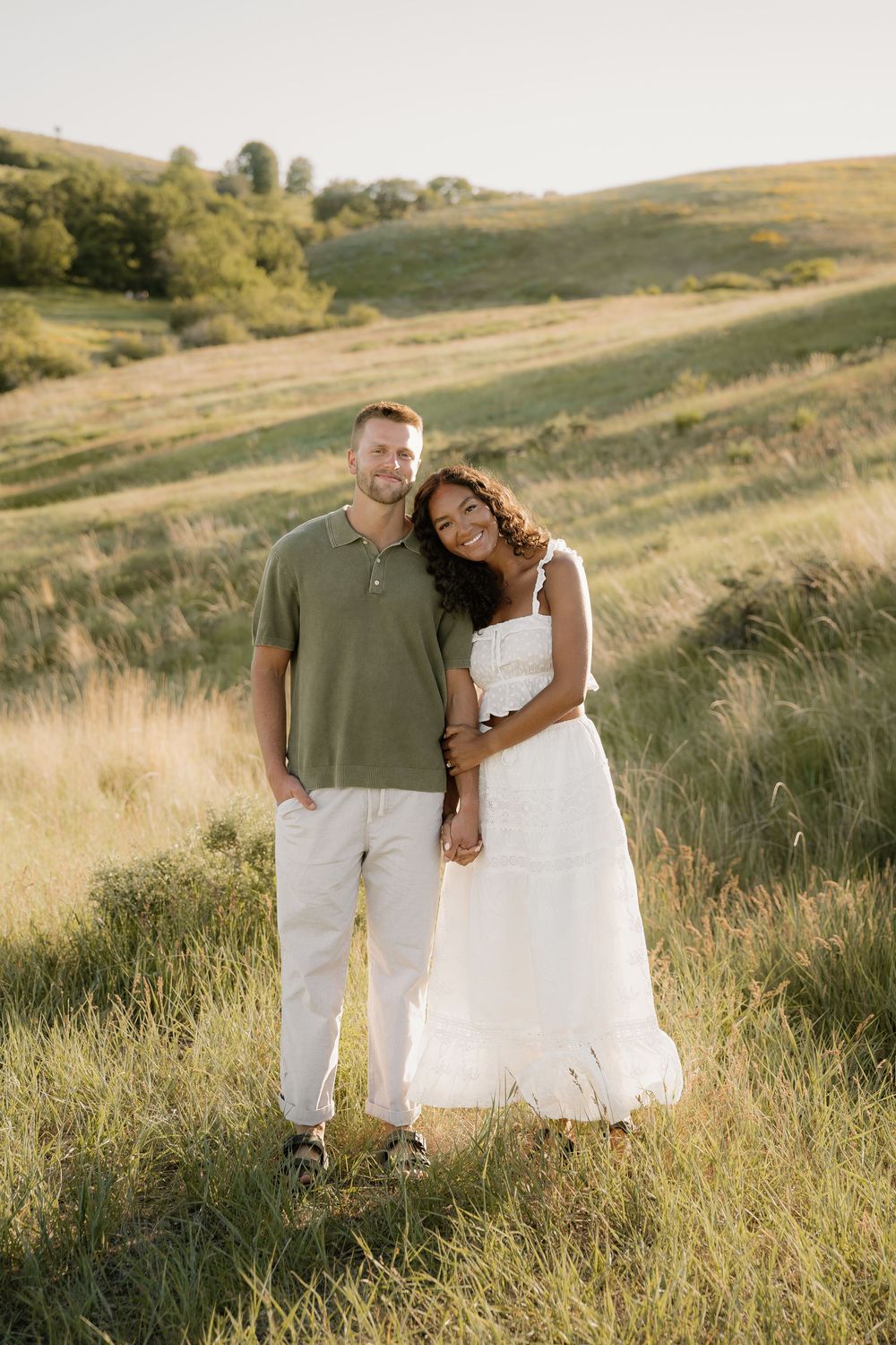 Couple poses casually in green field with rolling hills during golden hour photography session.