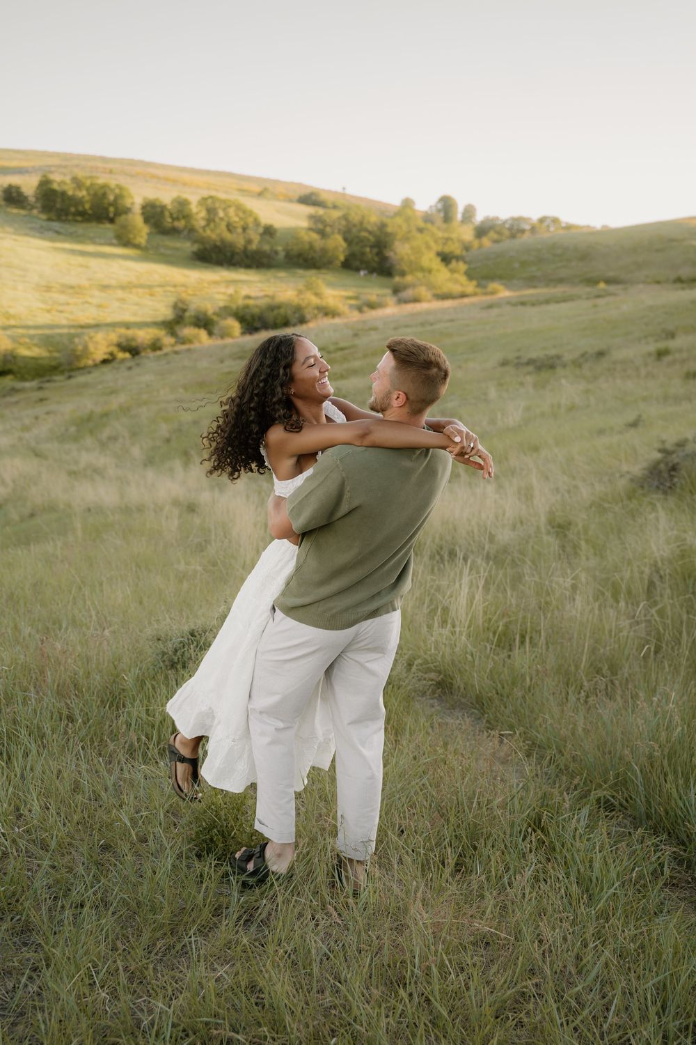 Romantic couple embracing in a golden sunlit meadow with rolling hills and trees in the background at sunset.