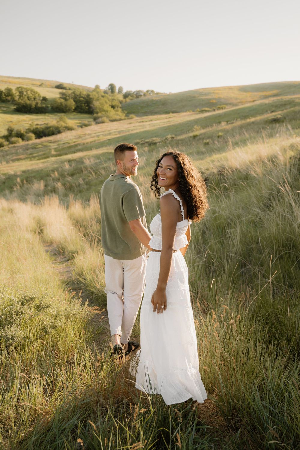 A romantic engagement photo of a couple standing in tall grass with rolling hills behind them.