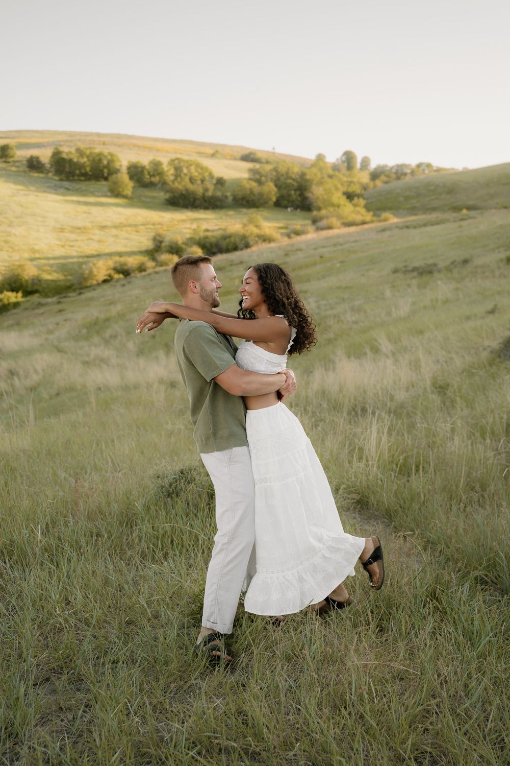 A couple shares a joyful embrace while standing in a hilltop meadow during golden hour.