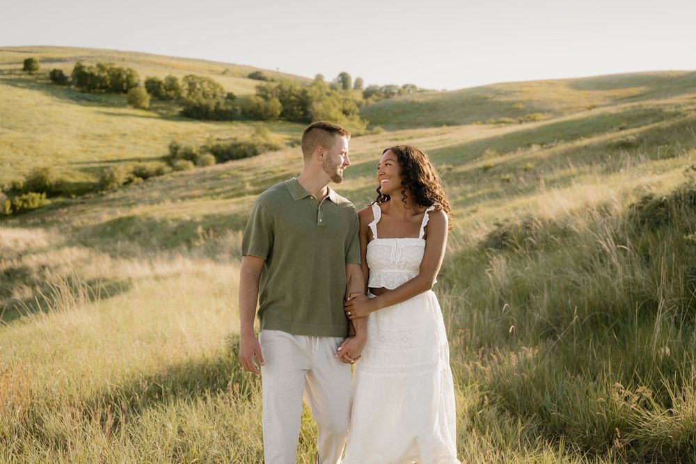 A couple in romantic wedding attire stands together on a rolling green hillside during golden hour.