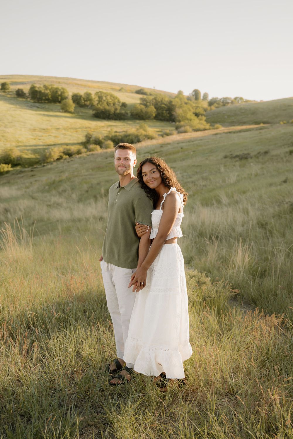 A couple stands embracing in a beautiful hillside meadow during sunset.