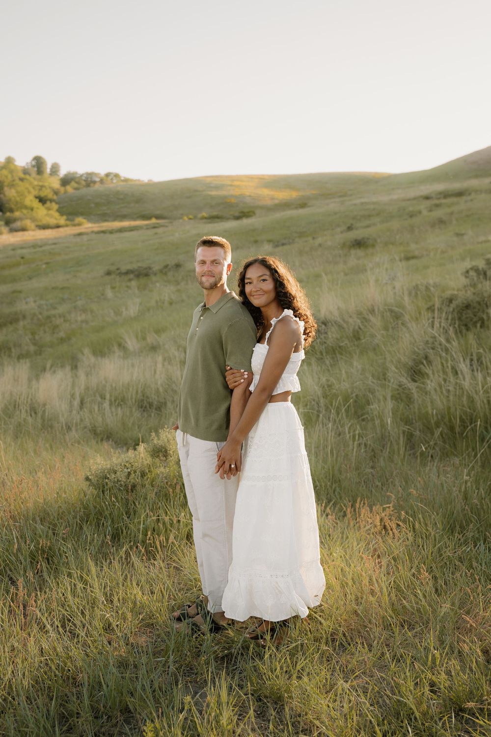 Couple poses together in casual outfits on grassy hillside during golden hour.