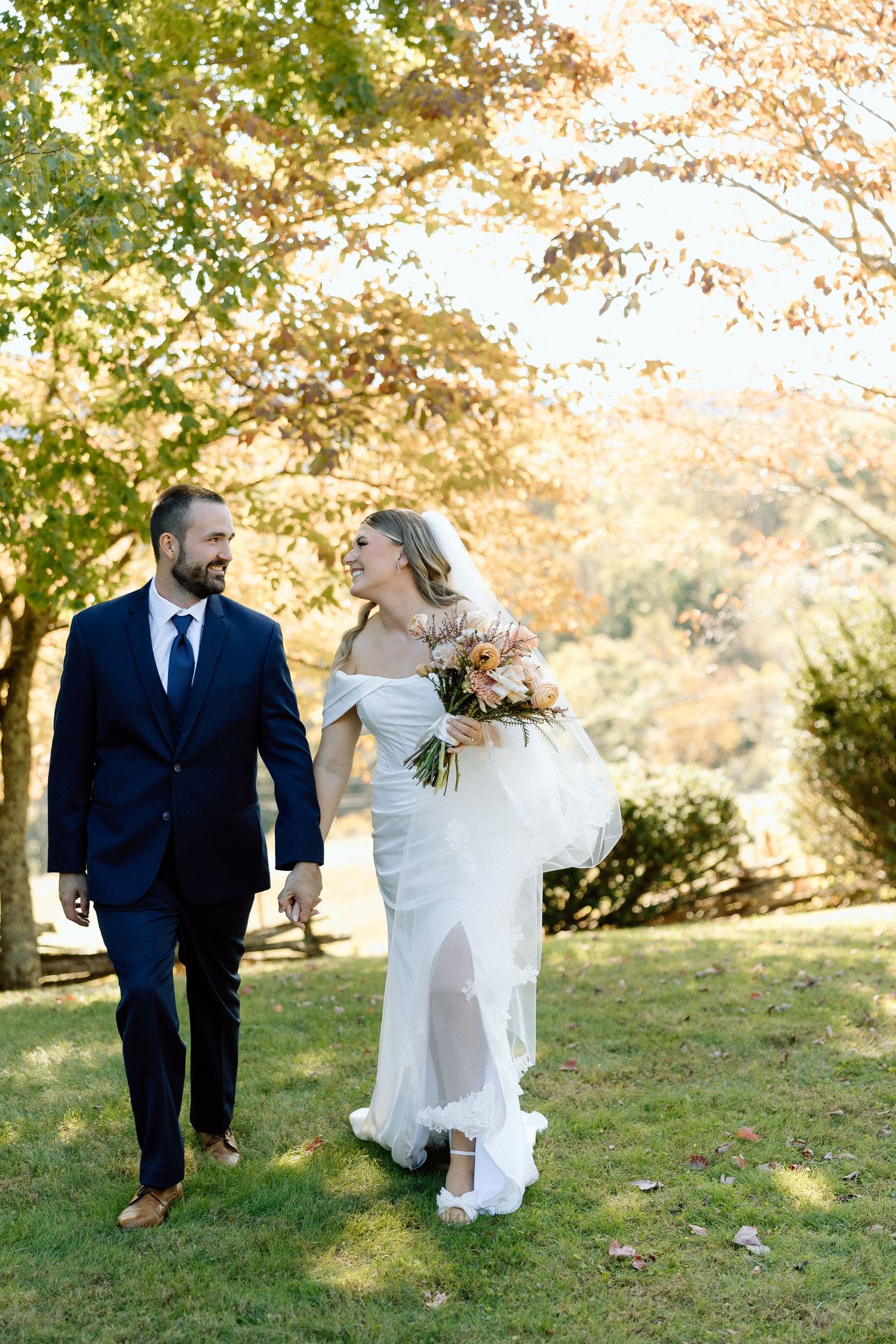 Beautiful fall wedding couple walks together on grassy lawn under golden autumn trees during sunset.