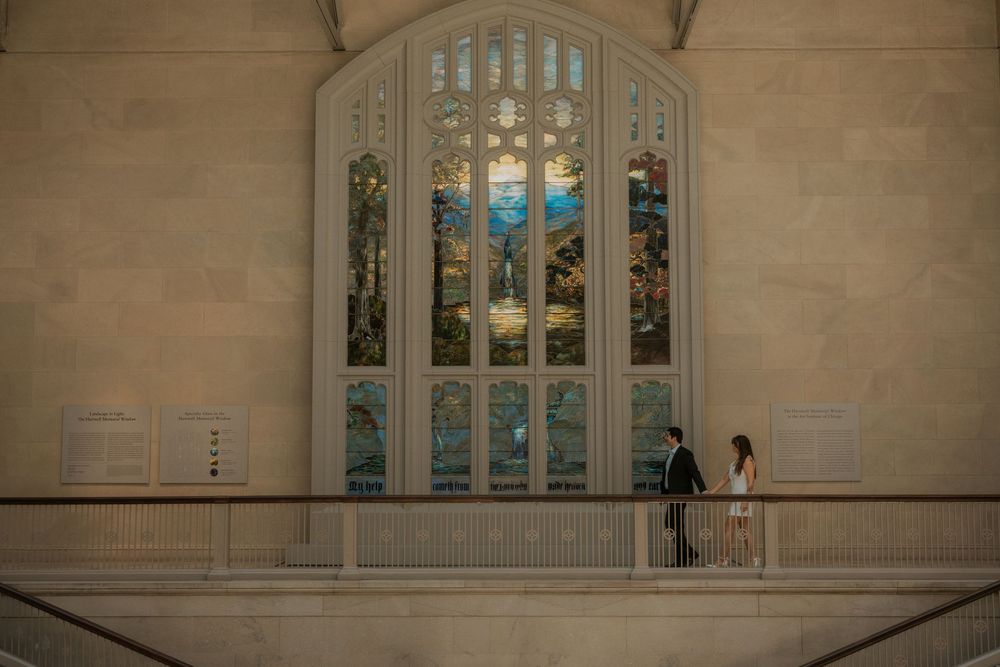 A couple stands silhouetted against a grand stained glass church window.
