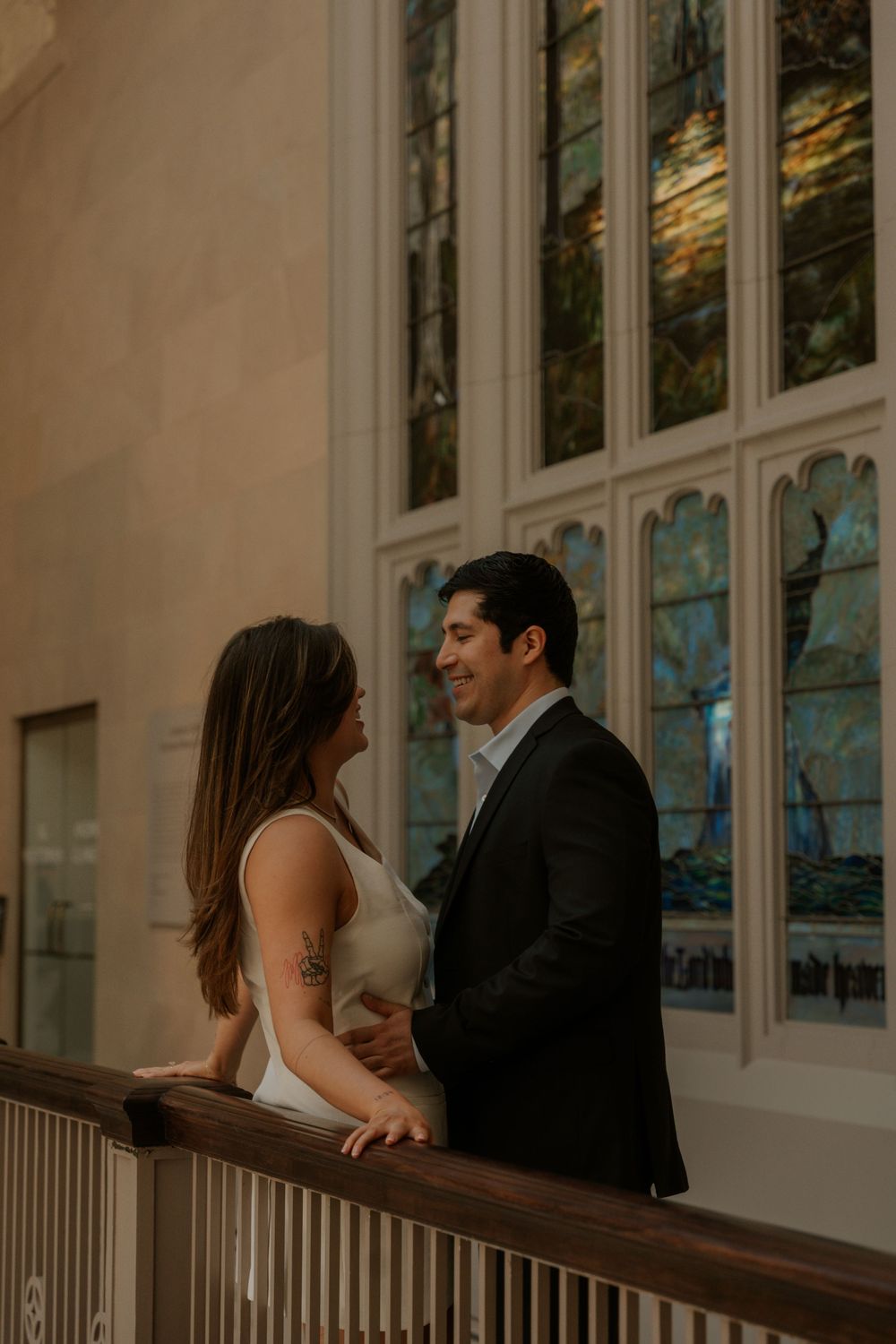 A romantic couple shares an intimate moment on a balcony with ornate windows in the background.