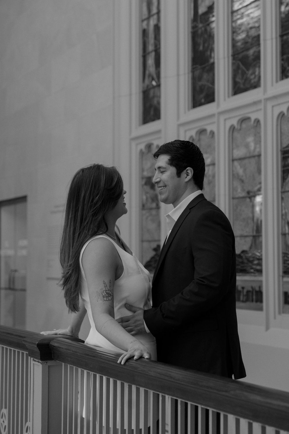 A romantic couple shares an intimate moment on an elegant balcony with ornate windows in the background.