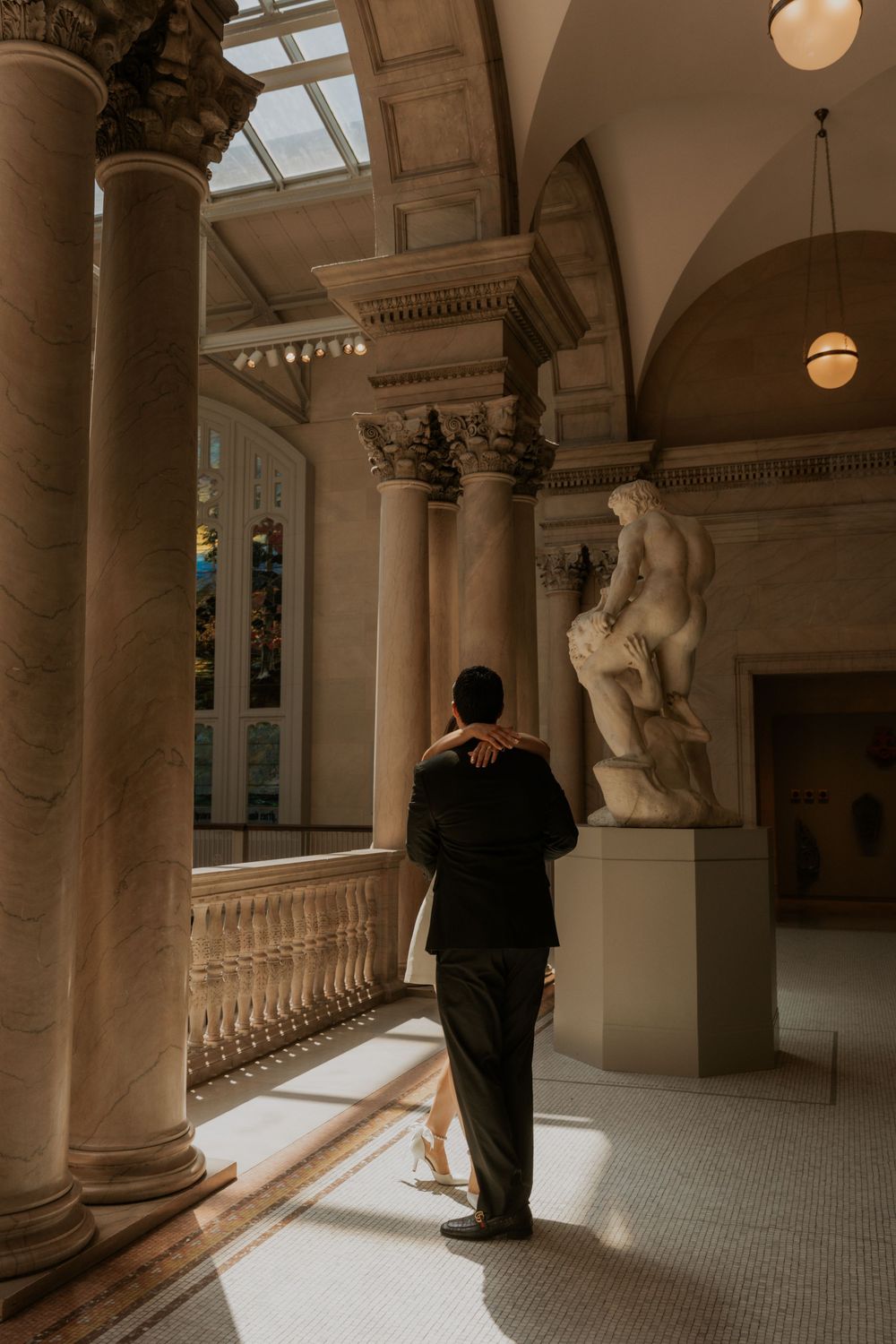 A figure stands in an elegant classical hallway with ornate columns and a marble statue illuminated by natural light.