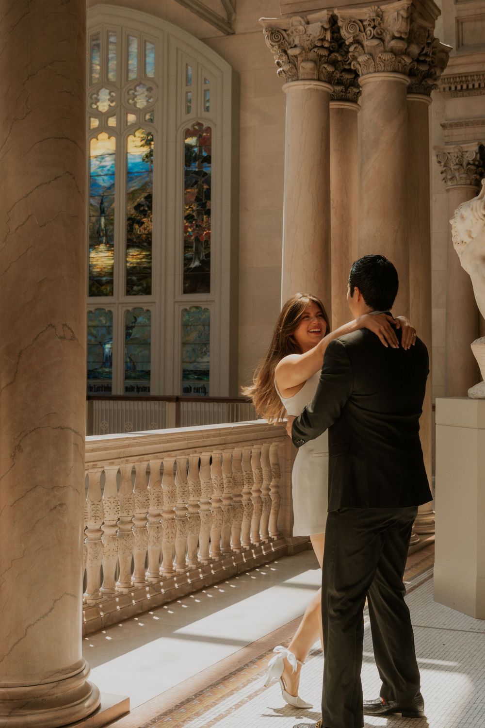 Couple dancing in elegant formal wear by classical stone columns and stained glass windows.