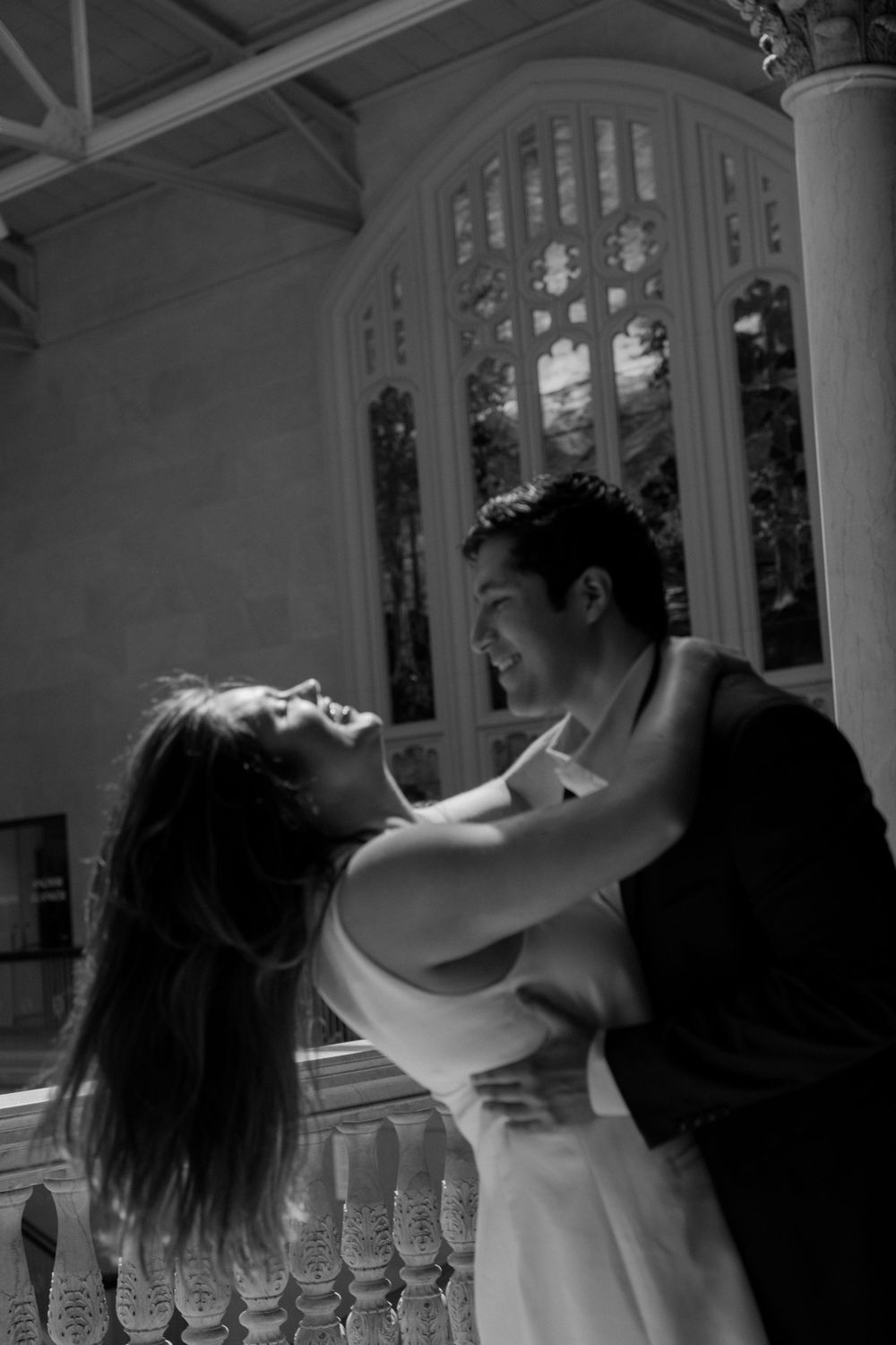 A romantic black and white portrait of a couple embracing intimately on a porch with ornate railing and window details.