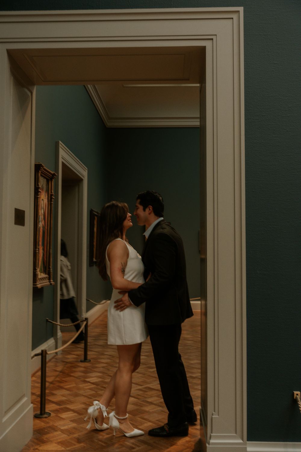 A couple embraces in a doorway of an art gallery with hardwood floors.