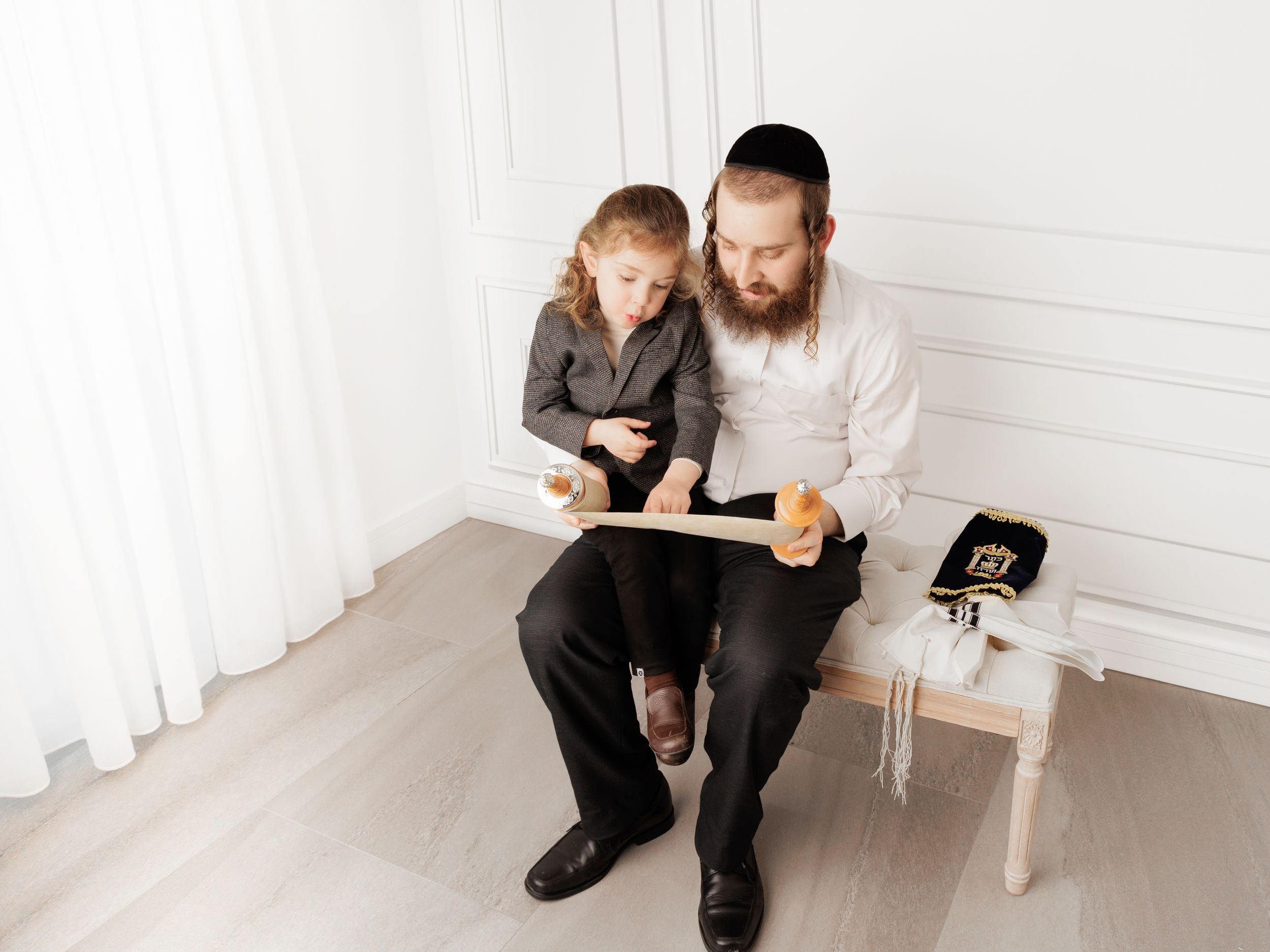 Adult and child sit together on floor reading a book in minimalist white room with curtains and small side table.
