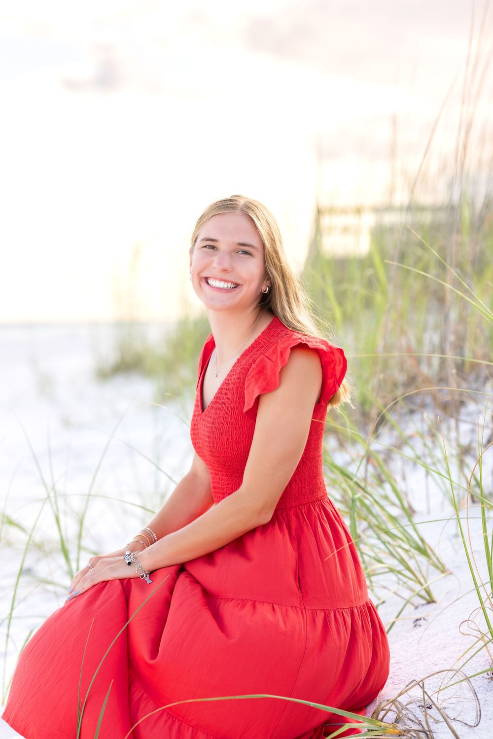 A smiling person in a red tiered dress sits among beach grass and sand dunes at sunset.
