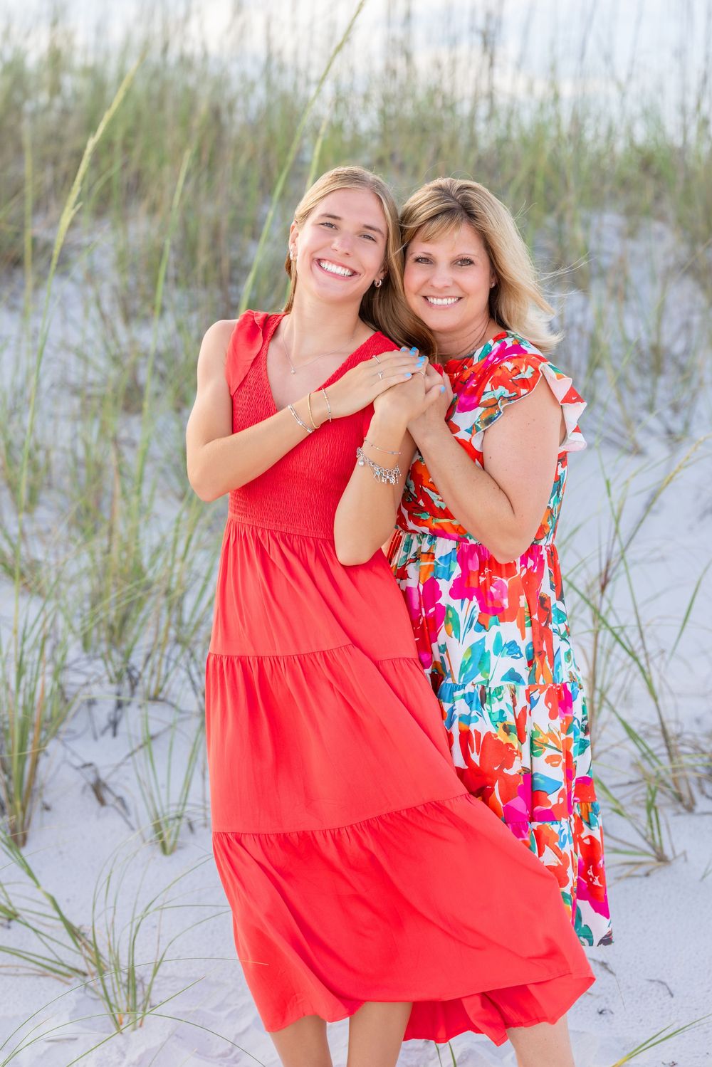 Two people wearing summer dresses pose together on a white sand beach with sea grass in the background.
