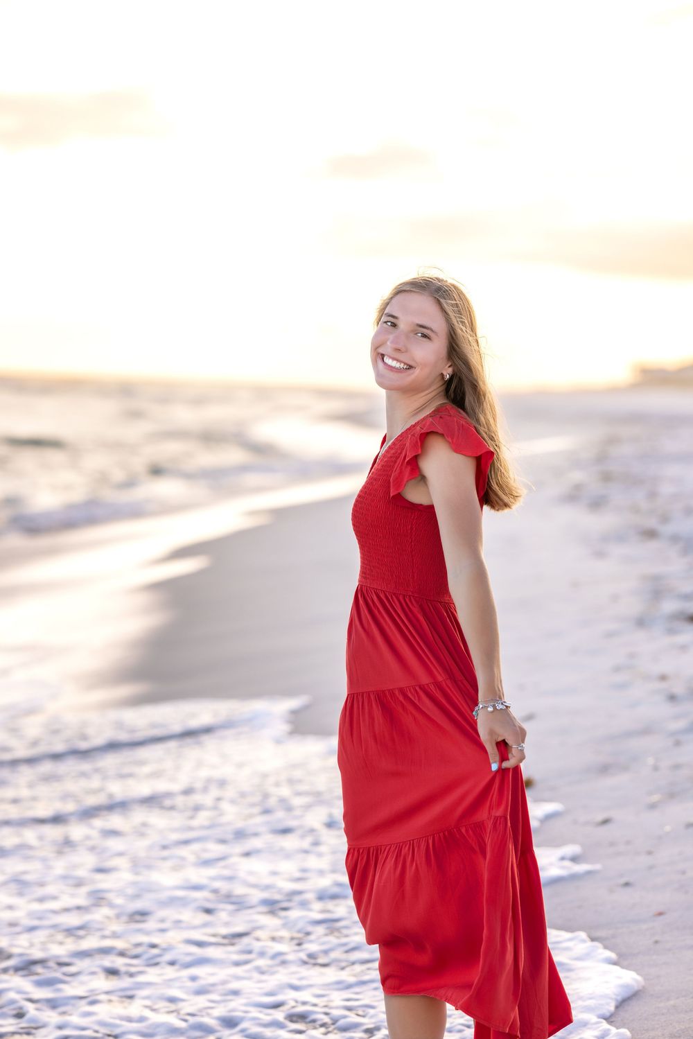 A person in a flowing red dress walks along a beach at sunset with their hair blowing in the breeze.