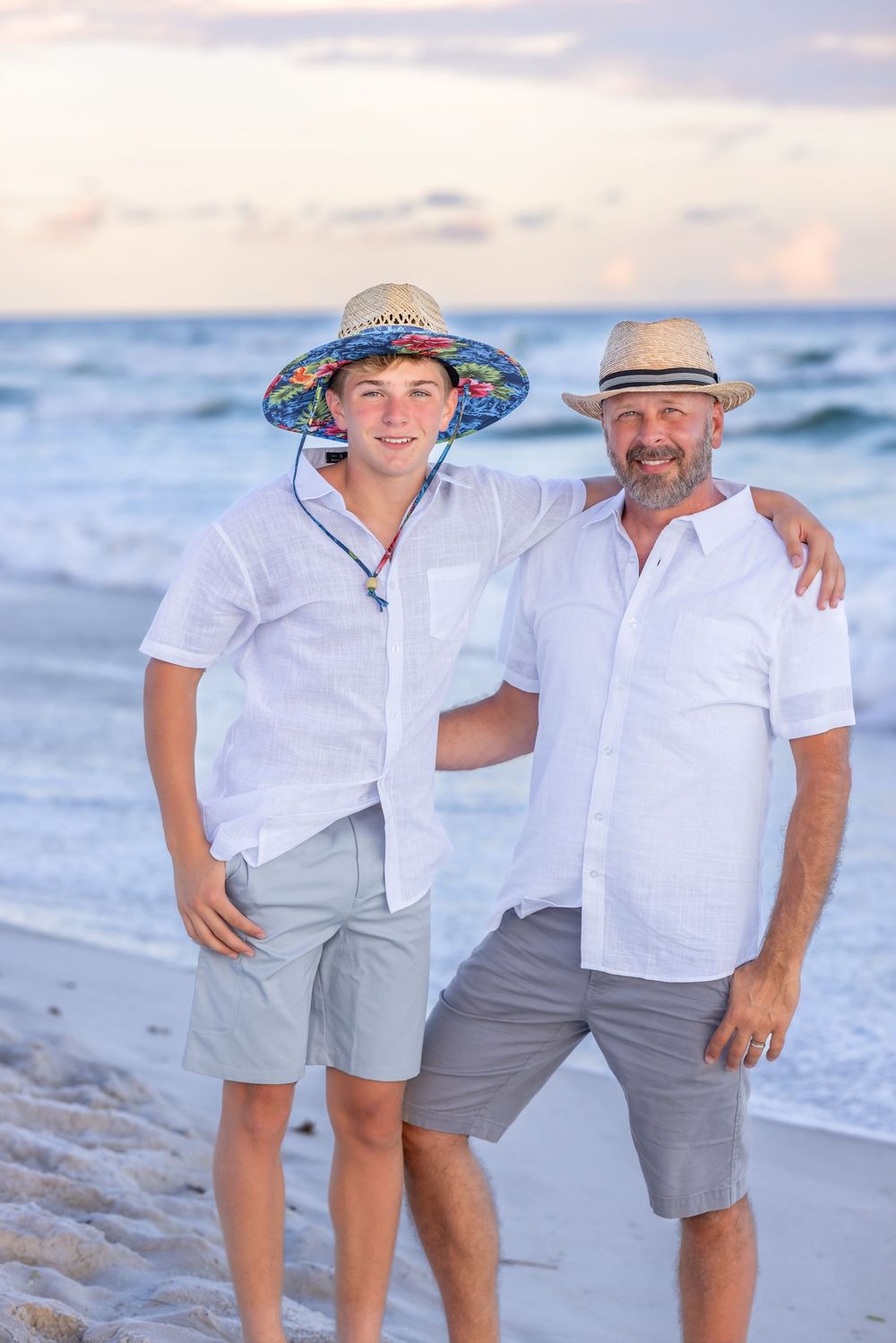 Two people in white shirts and gray shorts standing together on a beach at sunset wearing sun hats.