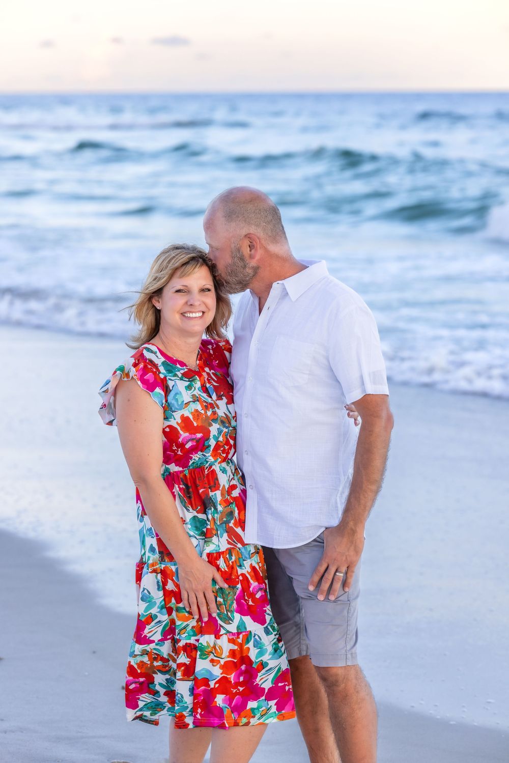 A couple stands on the beach at sunset wearing a vibrant red floral dress and white shirt against ocean waves.