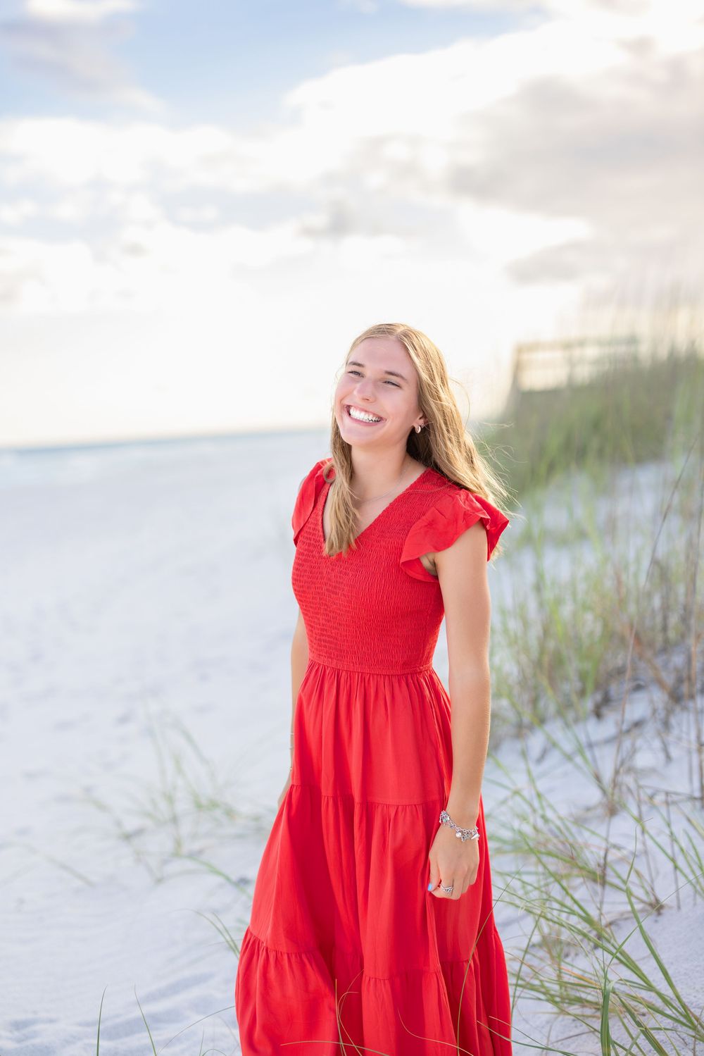 Young woman in flowing red tiered maxi dress smiles while standing on a sandy beach with dune grass and ocean view.