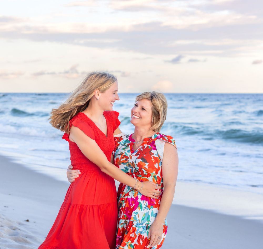 Two women in bright red and floral dresses share a joyful moment while walking along a beach at sunset.