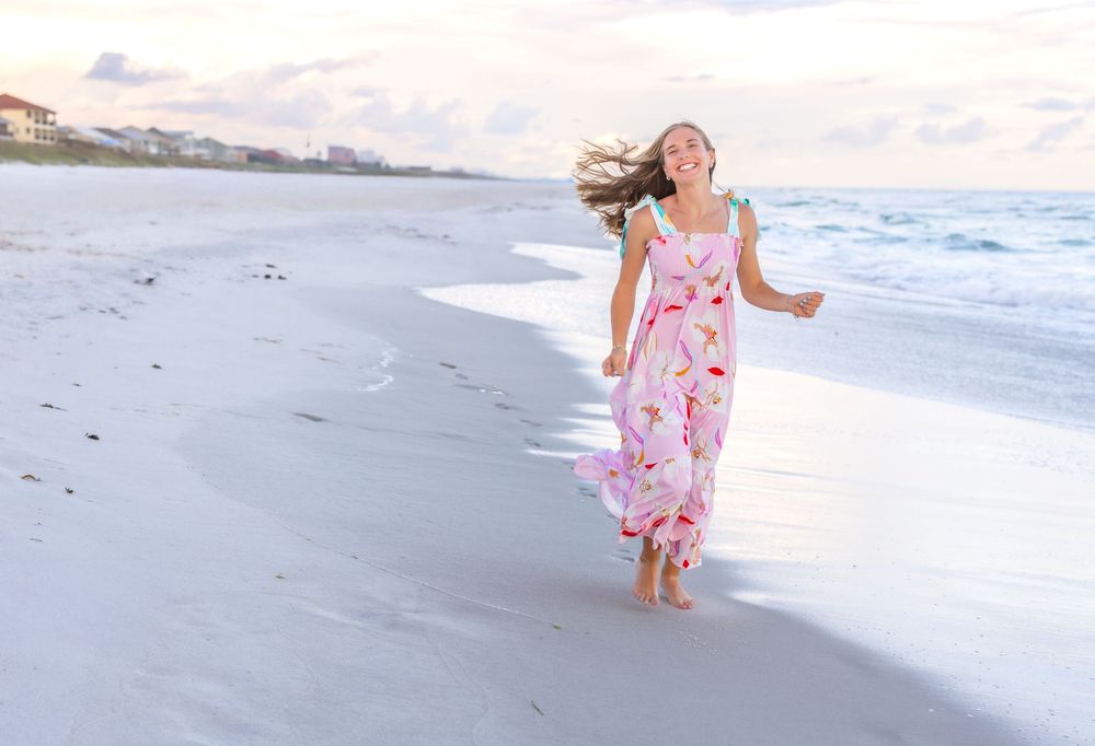 A person wearing a pink floral dress runs joyfully along an empty beach shoreline at sunset.