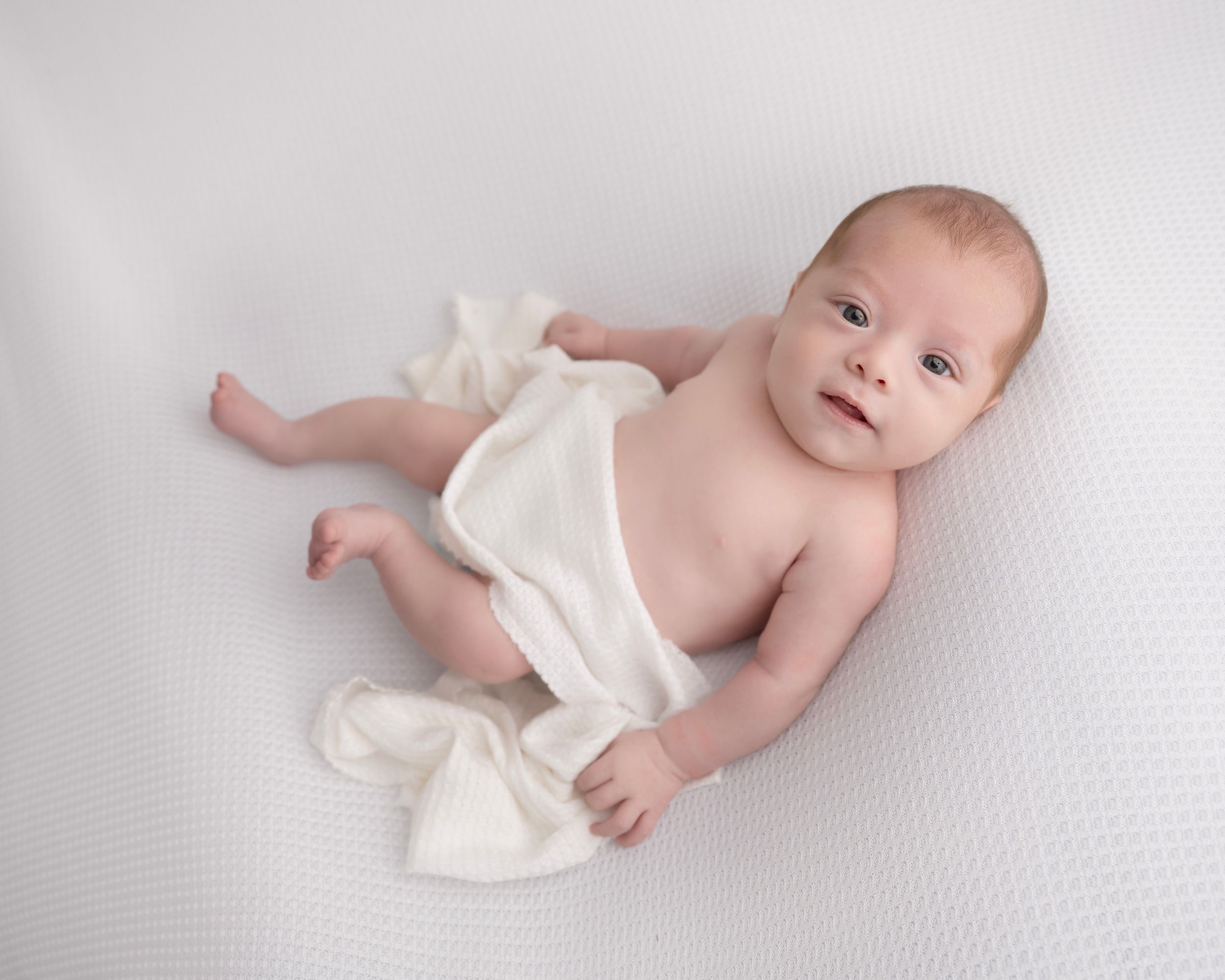 Small baby lying on white textured surface partially covered with soft cream-colored blanket.