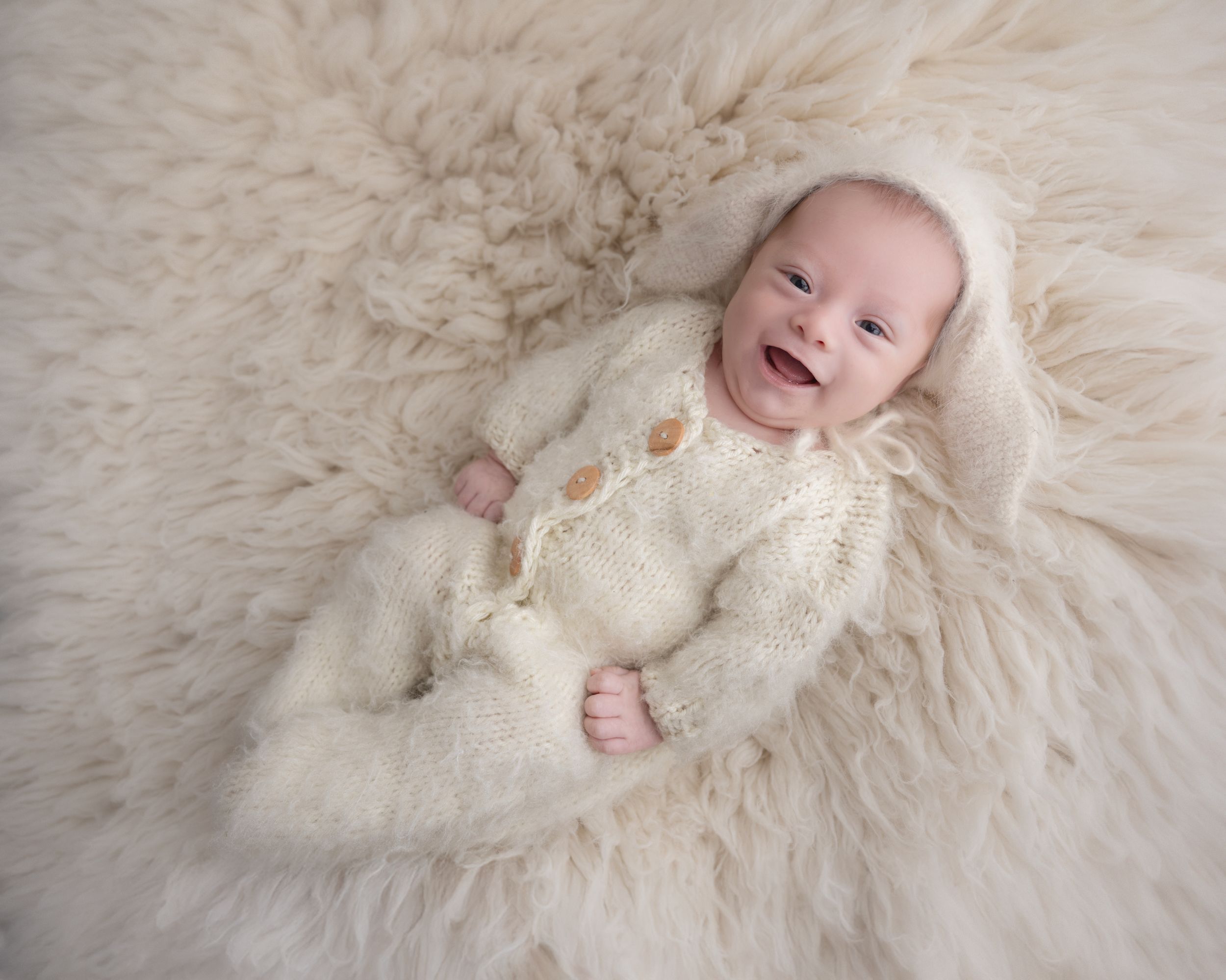 Smiling baby in white knit outfit lying on plush cream-colored fur blanket.