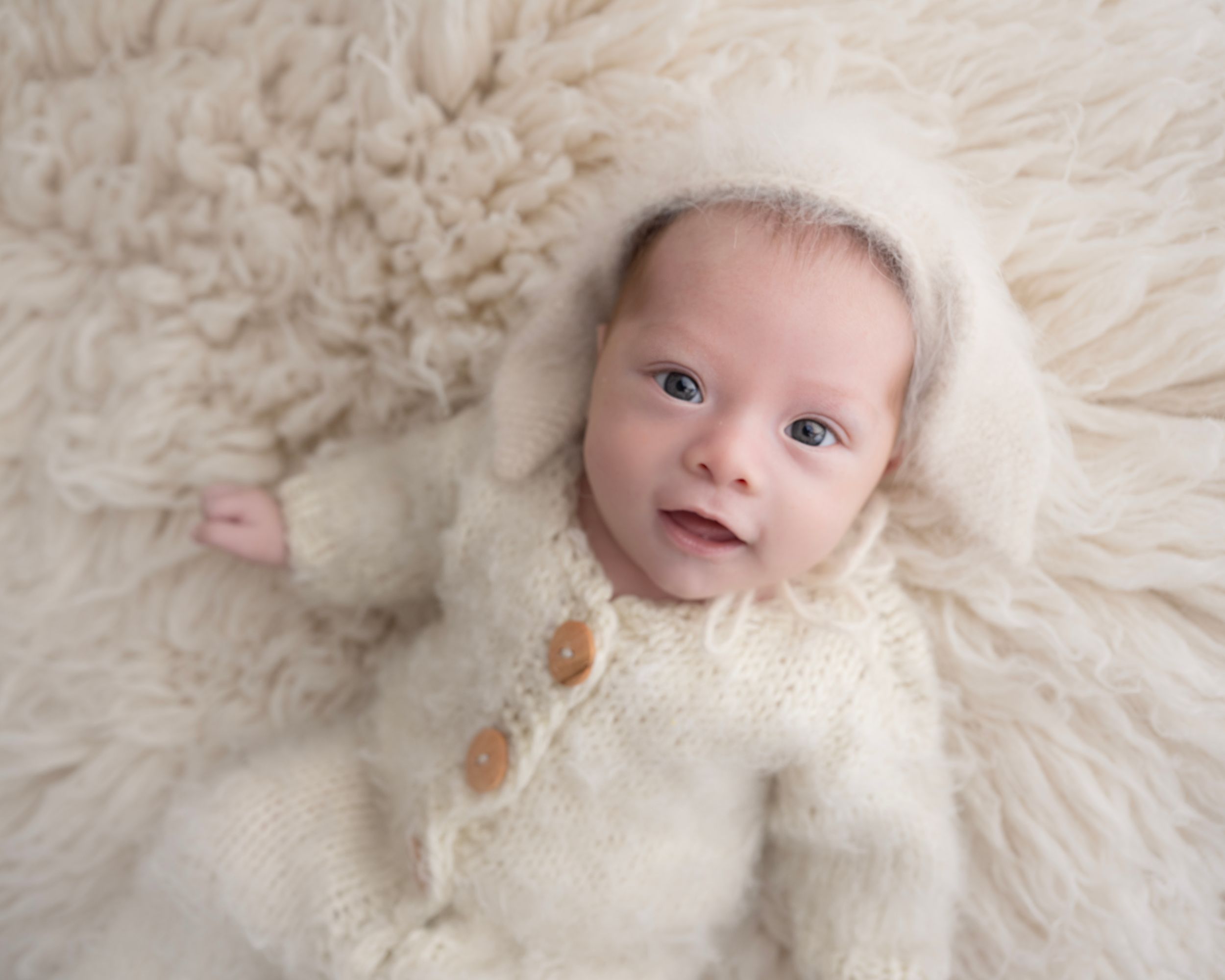 Newborn wearing cream colored outfit lying on soft white fur backdrop in studio setting.